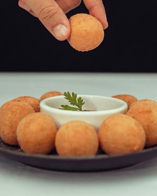 Mão pegando um bolinho de charque com banana; embaixo, outros bolinhos num prato escuro, em volta de um potinho de molho