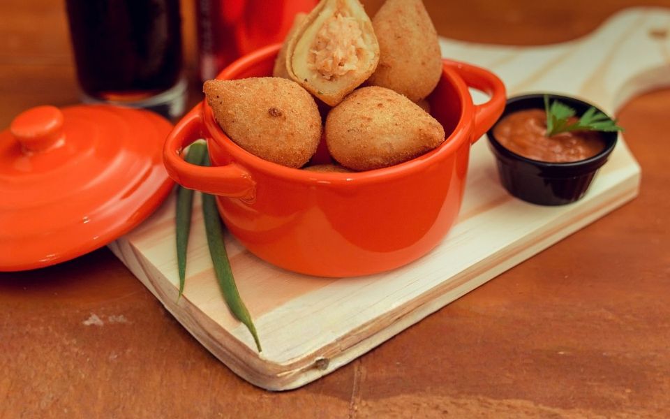 Coxinhas de frango numa panelinha laranja sobre uma tábua, uma delas aberta mostrando o recheio, com um potinho de molho ao lado