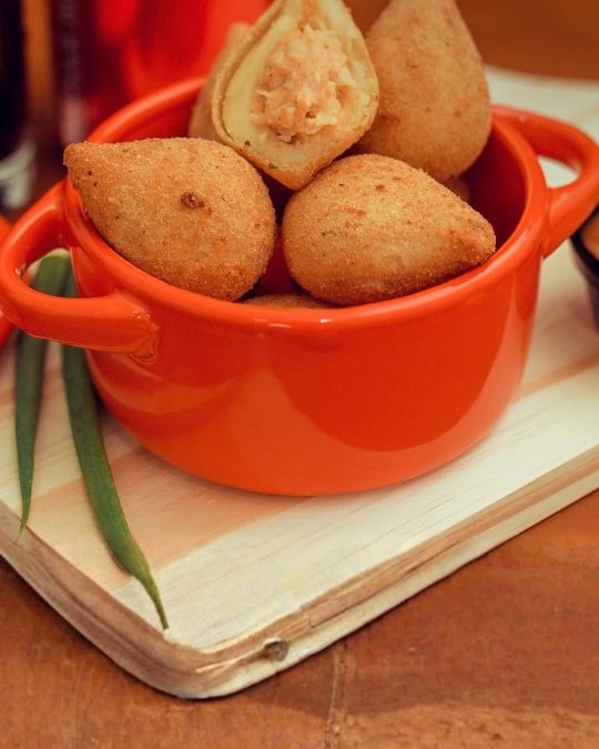Coxinhas de frango numa panelinha laranja sobre uma tábua, uma delas aberta mostrando o recheio